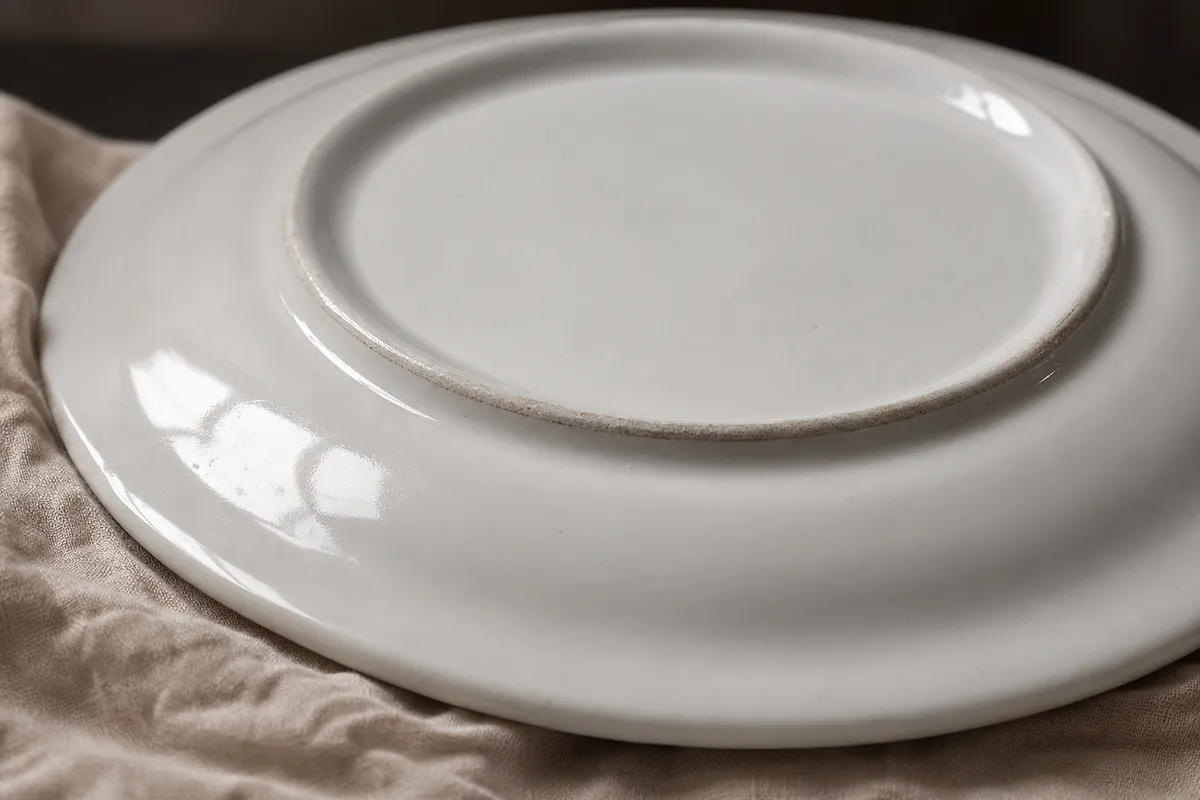 Revers d une assiette ancienne en porcelaine blanche posee sur un drap de lin, lumiere rasante revelant un tampon efface sous l email